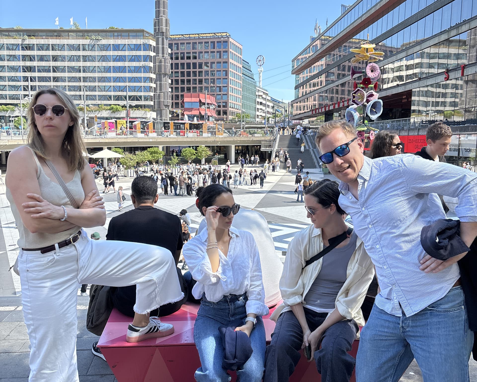 A group pausing on a busy sunlit city square, phones in hand, between scavenger hunt checkpoints.