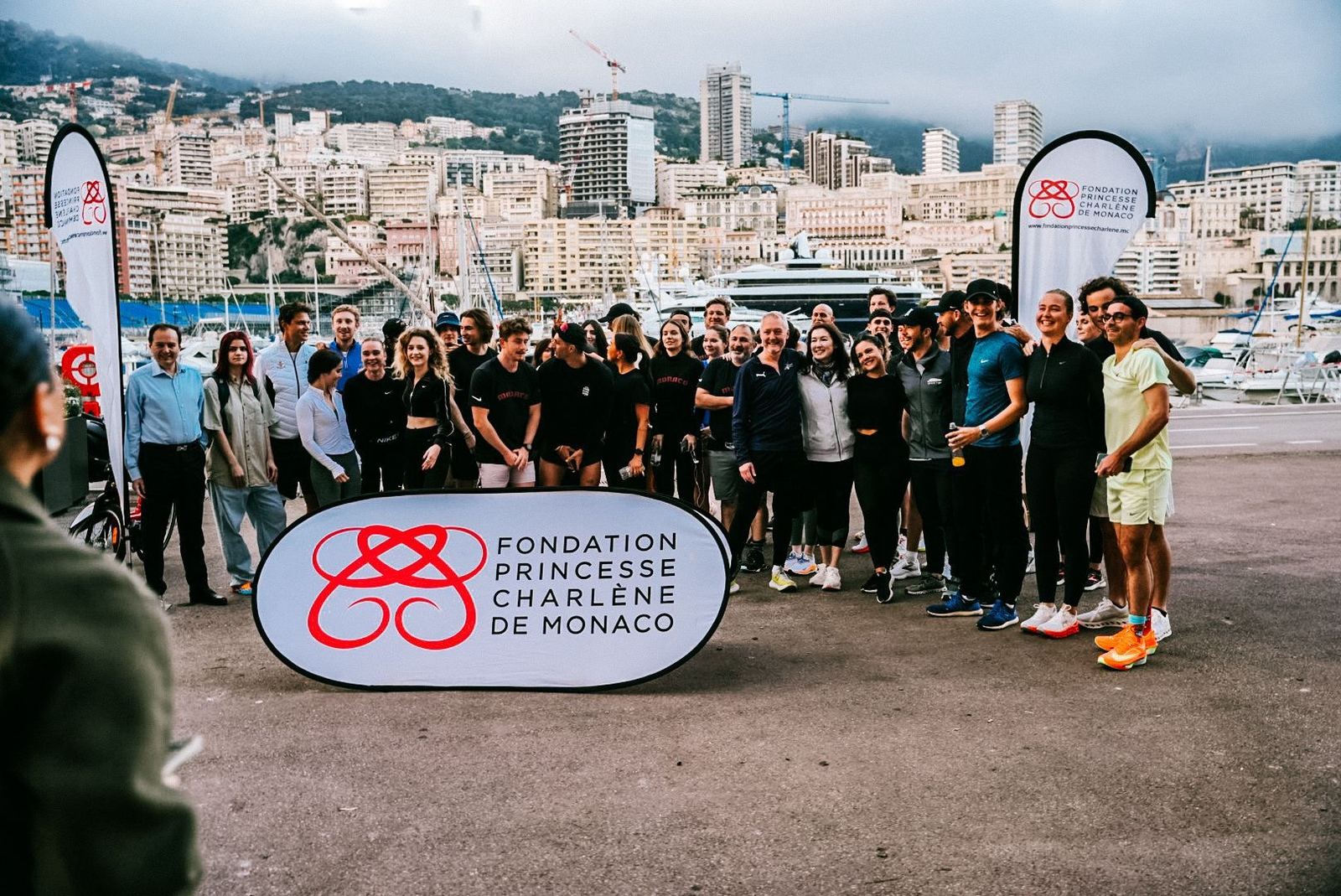 The Monaco participant group photographed beside the Fondation Princesse Charlène banner with the harbour and city skyline behind them.
